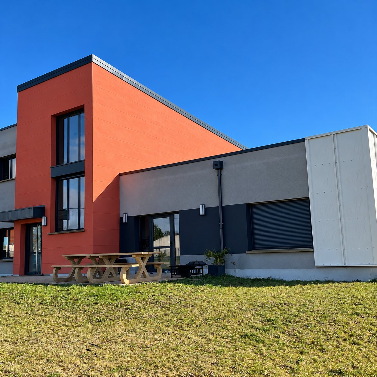 Façade d'un bâtiment moderne aux murs orange et gris sous un ciel bleu, avec une table de pique-nique en bois devant une pelouse.