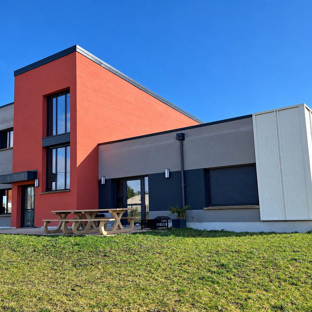 Façade d'un bâtiment moderne aux murs orange et gris sous un ciel bleu, avec une table de pique-nique en bois devant une pelouse.