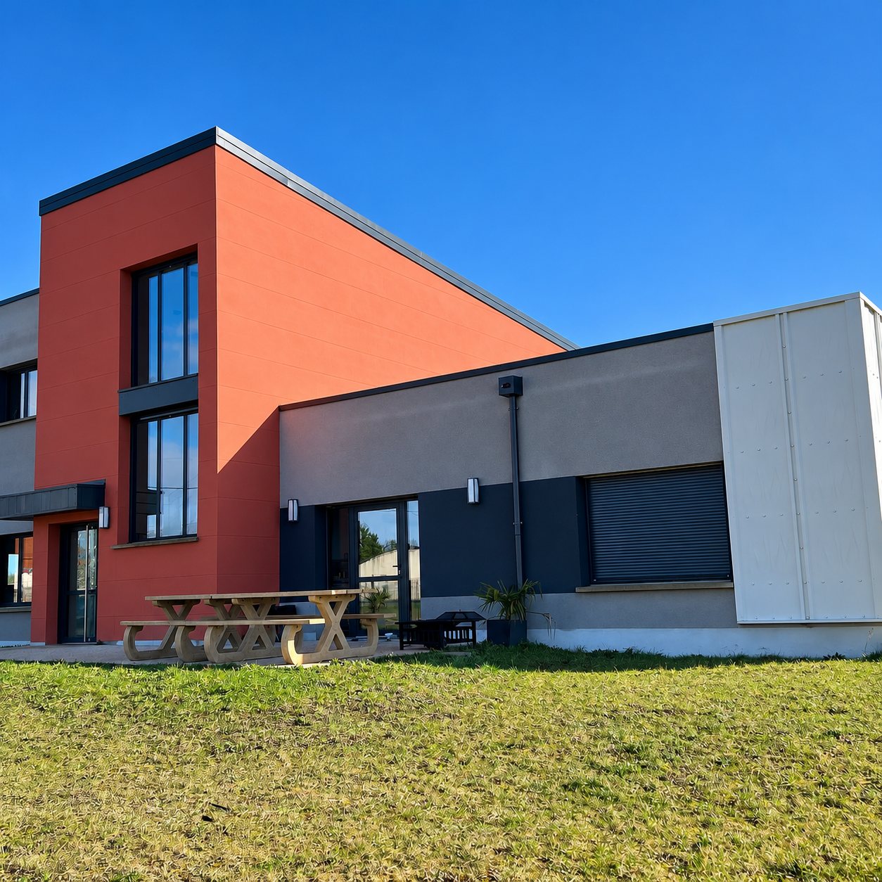 Façade d'un bâtiment moderne aux murs orange et gris sous un ciel bleu, avec une table de pique-nique en bois posée devant une pelouse.