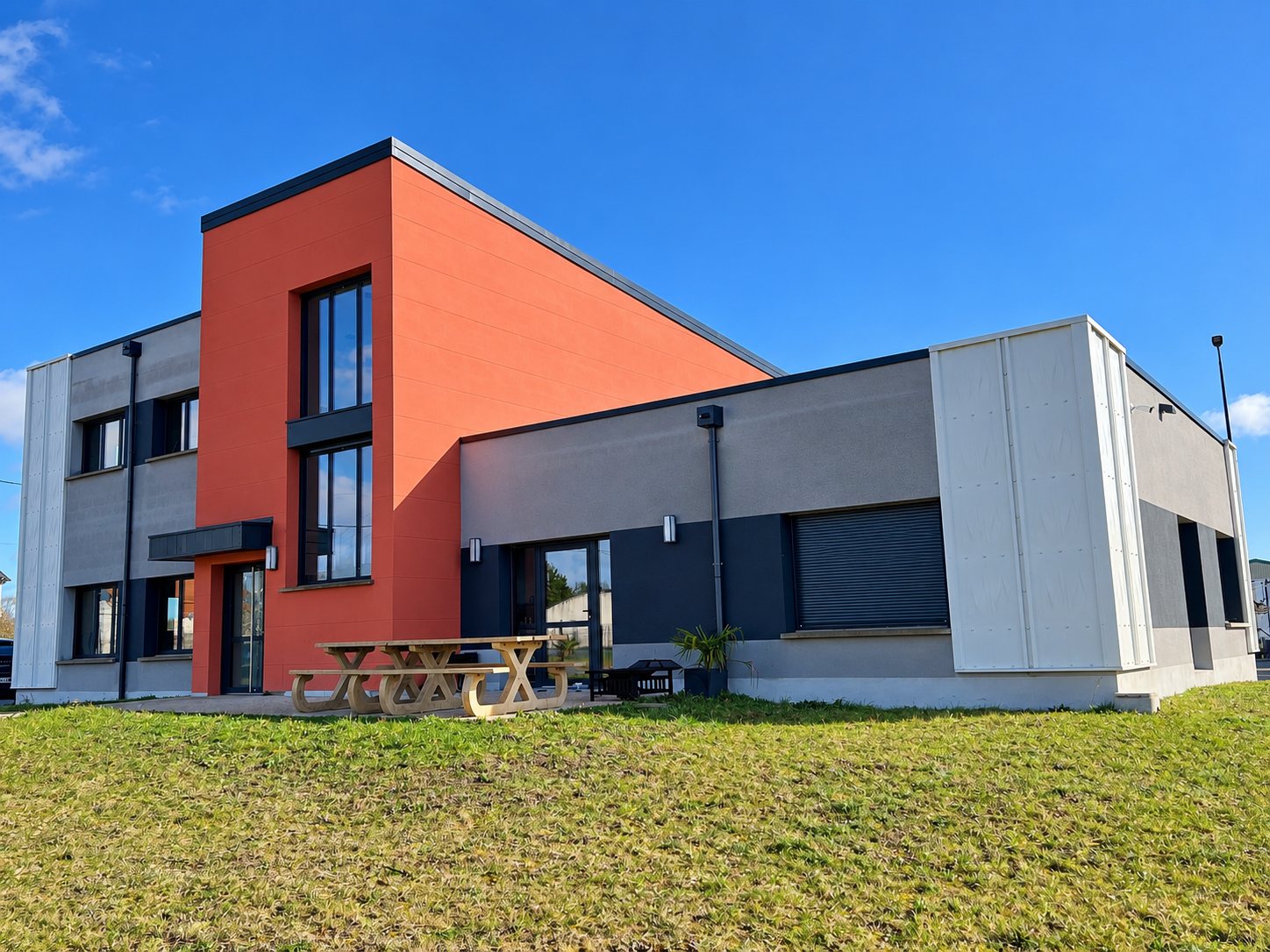 Façade d'un bâtiment moderne aux murs rouge brique et gris anthracite, avec une table de pique-nique en bois devant, sous un ciel bleu.