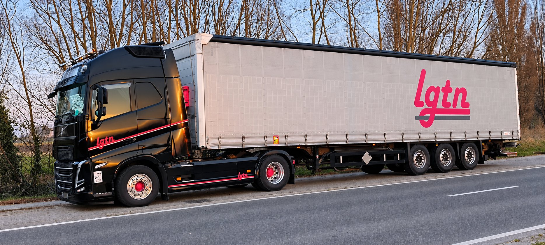 Un camion noir aux liserés rouges tracte une remorque à rideaux coulissants portant le logo lgtn, garé au bord d'une route au coucher du soleil.