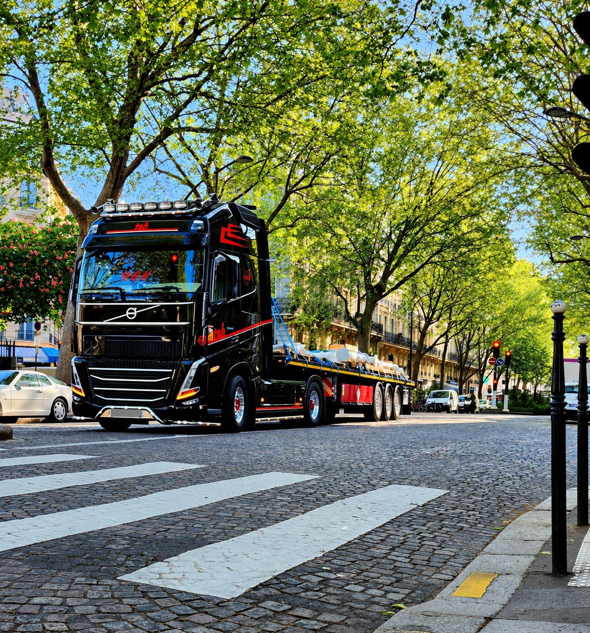 Un camion Volvo noir aux liserés rouges, équipé d'un plateau chargé, roule sur une avenue pavée bordée d'arbres feuillus près d'un passage piéton.