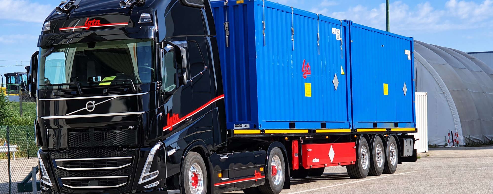 Un camion Volvo noir à liseré rouge tracte une semi-remorque chargée de deux conteneurs bleus, garé sur une aire goudronnée devant un grand abri en toile.