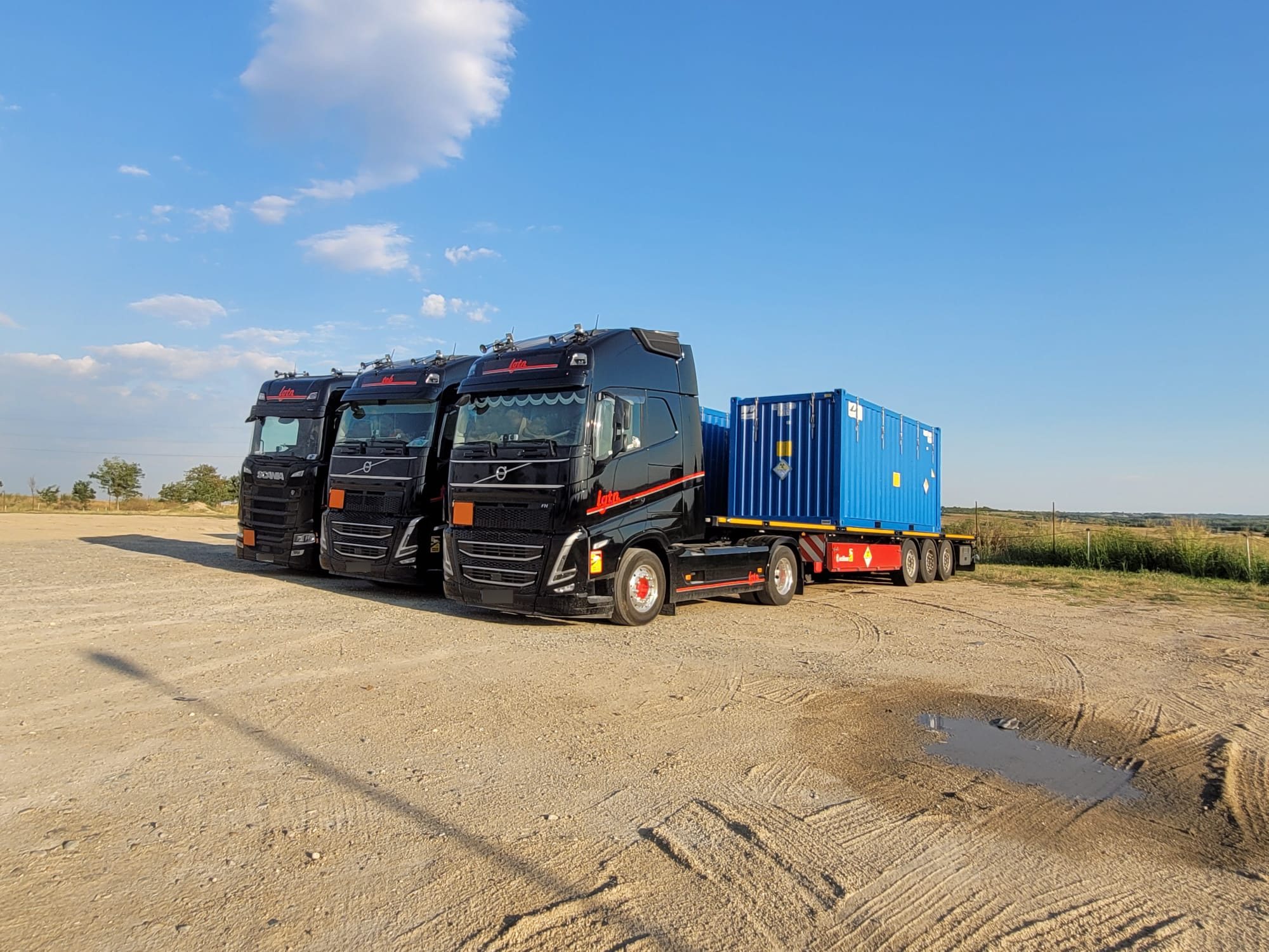 Trois camions noirs alignés sur un terrain de gravier sous un ciel bleu, le camion de droite transportant un conteneur bleu sur sa remorque.