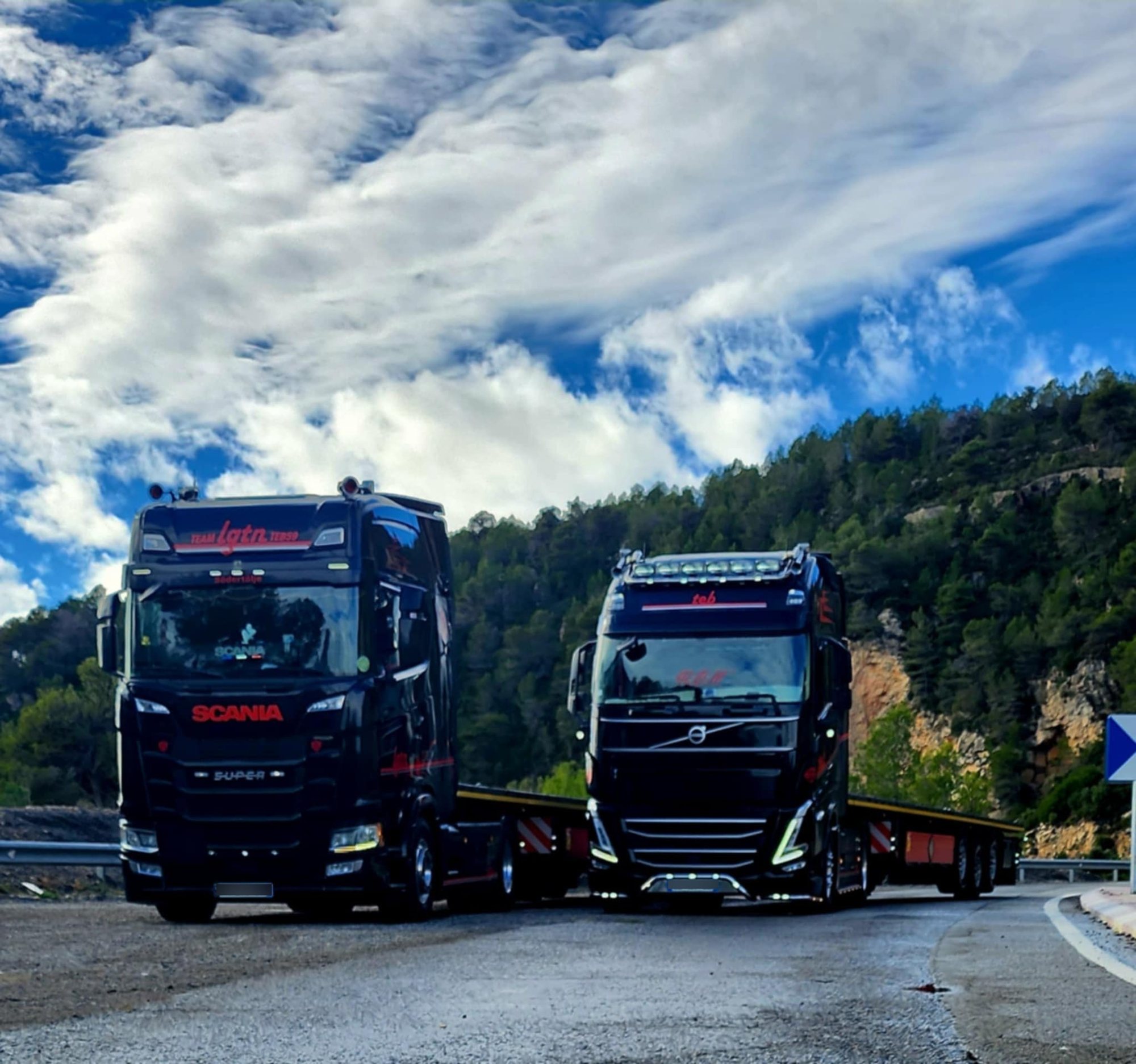 Deux camions noirs, un Scania et un Volvo, tractant des remorques plateaux, garés sur une route de montagne sous un ciel nuageux.