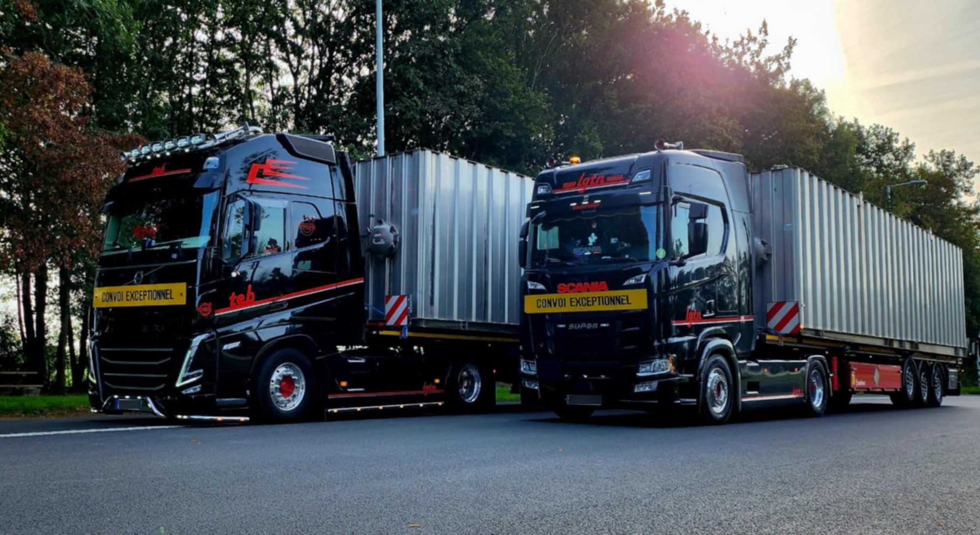 Deux camions noirs aux décors rouges, portant des panneaux jaunes « Convoi exceptionnel », sont garés côte à côte avec de grandes remorques métalliques sur une route bordée d'arbres.