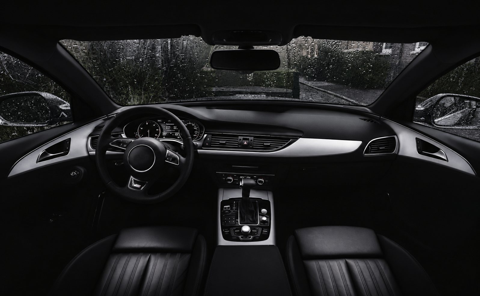 Photo en noir et blanc de l'intérieur d'une voiture, avec volant, tableau de bord, console centrale et sièges en cuir, derrière un pare-brise couvert de gouttes de pluie.